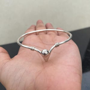 May include: A silver-toned metal bangle bracelet featuring a V-shaped centre point adorned with a smooth, round sphere. The band includes decorative wire-wrapped detailing on both sides of the central sphere, creating a structured, open-ended design.