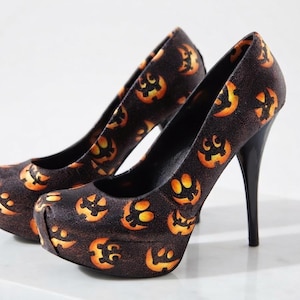 May include: A pair of high-heeled platform pumps featuring a dark, textured black base covered in a repeating pattern of orange and yellow glowing jack-o'-lantern faces. The shoes have a rounded toe, a thick platform sole, and a slim black stiletto heel.