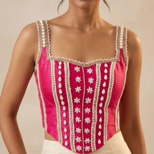 May include: A sleeveless fuchsia corset top featuring a square neckline and a pointed hem. The bodice is decorated with vertical rows of white pearl beads, small white floral appliqués, and gold-toned trim along the straps and seams.