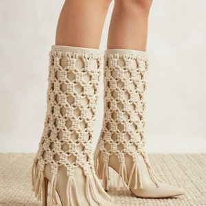 May include: A pair of cream-colored mid-calf boots featuring a pointed toe and a high stiletto heel. The upper portion of the boots is covered in a decorative macramé cord overlay with a knotted lattice pattern that extends into fringe at the base.