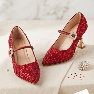 May include: A pair of red pointed-toe pumps covered in small red crystals. Each shoe features a thin satin strap with a square crystal-encrusted buckle and a gold-toned flared heel. The letter R is near the left shoe and L near the right.