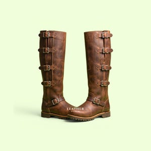 Puede incluir: Un par de botas altas de cuero marrón con cuatro hebillas plateadas verticales en el lado exterior de cada caña y una hebilla adicional en la zona del tobillo. Las botas tienen una suela gruesa y el texto LEATHER PUNCH.