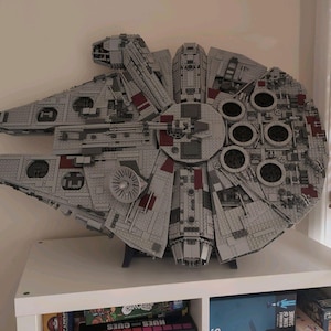 Könnte beinhalten: Ein großes, detailliertes Modell des Millennium Falcon Raumschiffs, das aus grauen Kunststoff-Bausteinen gefertigt ist. Die Struktur verfügt über einen kreisförmigen Hauptkörper, ein Cockpit an der Seite und komplexe mechanische Details.
