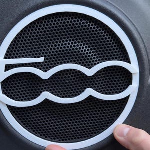 Puede incluir: Cubierta de altavoz blanca impresa en 3D con el logotipo del Fiat 500. La cubierta circular tiene un respaldo de malla y está colocada sobre una superficie gris oscuro. Un dedo toca el borde de la cubierta.