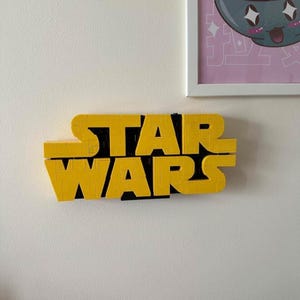 Op de afbeelding: Een wandbord met de tekst STAR WARS, opgebouwd uit gele in elkaar grijpende plastic bouwstenen. De letters zijn in twee rijen op een zwarte achtergrond geplaatst, wat zorgt voor een driedimensionaal en blokkerig uiterlijk.