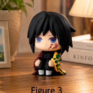 Puede incluir: Peque&ntilde;a figura de anime con cabello negro, ojos azules y una marca roja en la cara. La figura lleva un atuendo negro con una capa roja y sostiene un objeto amarillo y verde. El texto "Figure 3" est&aacute; en la parte inferior.
