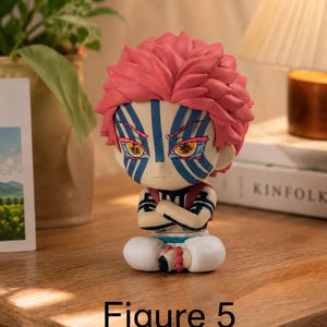 Puede incluir: Figura detallada de un personaje con pelo rosa puntiagudo, marcas faciales azules y blancas y brazos cruzados. La figura est&aacute; sentada con pantalones blancos y una pulsera de cuentas rojas. El texto "Figure 5" est&aacute; en la base.