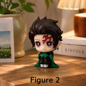 Puede incluir: Una peque&ntilde;a figura de un personaje con cabello oscuro y un dise&ntilde;o rojo y negro en la frente. La figura viste un atuendo a cuadros verdes y negros. El texto "Figure 2" est&aacute; en la parte inferior de la imagen. La figura est&aacute; sobre una superficie de madera.
