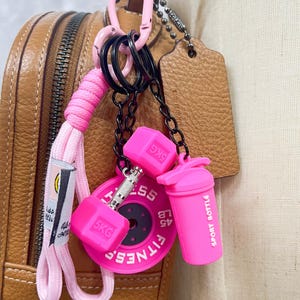 Puede incluir: Un conjunto de llaveros que incluye un cordón trenzado rosa, un dije de mancuerna rosa con la etiqueta 5KG, un dije de disco de peso circular rosa con la etiqueta FITNESS 45 LB y un dije de botella deportiva rosa, unidos a un mosquetón rosa.