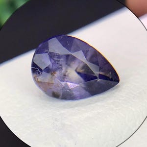 May include: A pear-shaped faceted gemstone with a deep violet and blue hue, featuring internal inclusions and a polished surface that reflects light across its geometric cut facets, showcasing a mix of dark and light purple colour variations.
