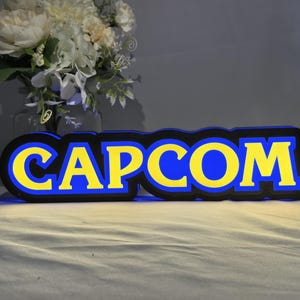 May include: A rectangular, illuminated sign featuring the word CAPCOM in bold, yellow, block-style lettering. The text is outlined in a thick blue border, which is further encased in a black frame. The sign emits a soft blue glow.