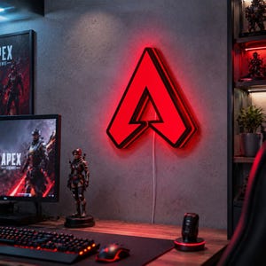 以下が含まれることがあります： 中央に切り抜きがあるスタイリッシュな「A」の形をした、Apex Legendsの赤いロゴの壁掛けネオンサイン。このサインは明るい赤色の光を放ち、灰色の壁に沿って垂直に垂れ下がった白い電源コードに接続されています。
