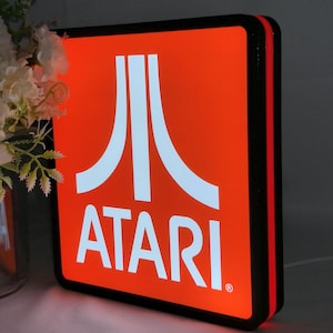 May include: A square, illuminated Atari sign featuring a bright orange background with the white Atari logo and text centered on the front. The sign has a thick black border and a thin red accent strip along the side edge of the frame.