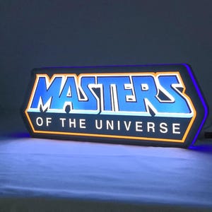 May include: A rectangular LED sign featuring the text MASTERS OF THE UNIVERSE in a bold, stylized blue font with a white outline. The sign is illuminated with a bright blue glow, framed by a thin orange border, and includes a black power cord.