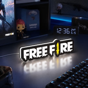 以下が含まれることがあります： 白い文字でFREE FIREと書かれた長方形のLEDサイン。文字のIの部分は黄色いナイフのグラフィックに置き換わっています。サインには黒い縁取りがあり、背面から明るい白色の光を放ち、下の暗い表面に反射しています。