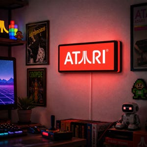 Può includere: Un'insegna rettangolare da parete con il logo Atari in caratteri bianchi su uno sfondo rosso vibrante. L'insegna è illuminata e proietta un bagliore rosso sulla parete circostante. È posizionata sopra una scrivania con accessori da gioco.