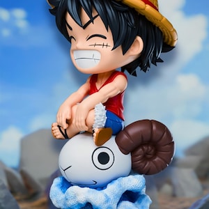 Puede incluir: Una figura estilizada de un personaje con cabello negro, sombrero de paja, camiseta roja sin mangas y pantalones cortos azules, sentado sobre una criatura blanca con forma de carnero y cuernos marrones, sobre una base de olas azules.