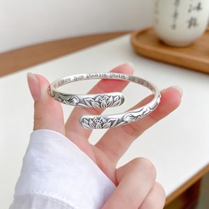 May include: A silver open cuff bracelet featuring embossed lotus flower motifs on both ends and Sanskrit text engraved along the inner band. The metal has a matte, oxidized finish that highlights the intricate floral details and lettering.