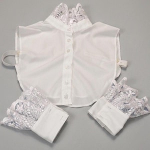 May include: A white detachable shirt collar and matching pair of cuffs. The collar features a high neck with intricate white lace trim and a front button placket. The two cuffs also feature matching white lace trim and button closures.