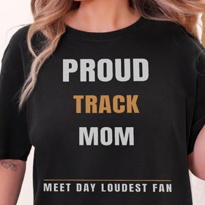 May include: A black short-sleeved t-shirt featuring the text PROUD in light gray, TRACK in gold, and MOM in light gray. Below this, a horizontal gold line sits above the text MEET DAY LOUDEST FAN in light gray, centered on the chest area.