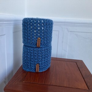 May include: Two blue crocheted toilet paper roll covers with brown leather labels. The covers are stacked on top of each other on a wooden surface.