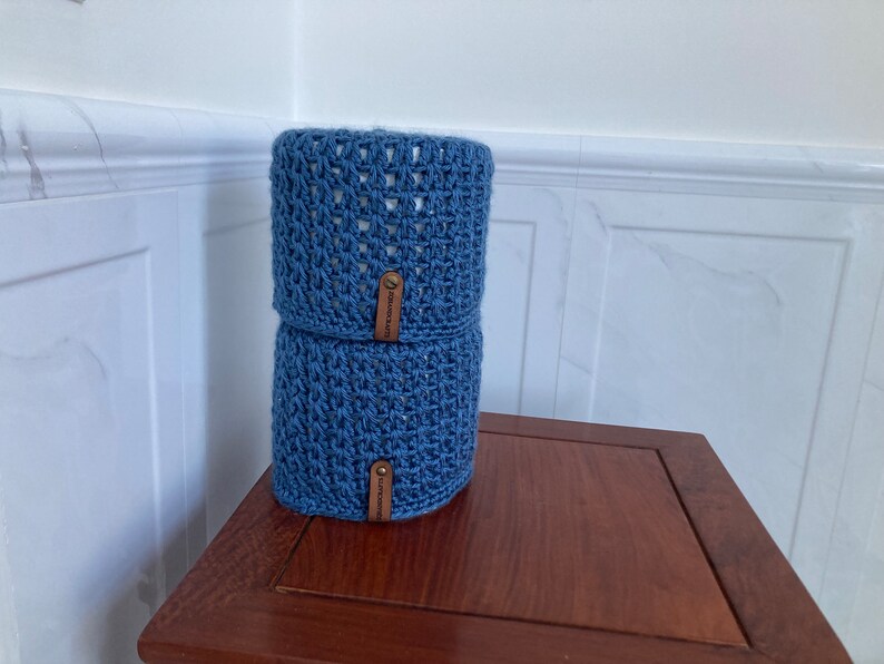 May include: Two blue crocheted toilet paper roll covers with brown leather labels. The covers are stacked on top of each other on a wooden surface.