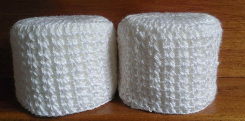 May include: Two white crocheted toilet paper roll covers. The covers are made with a simple stitch pattern and have a slightly textured look.