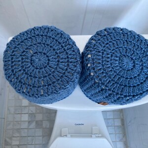 May include: Two blue crocheted toilet paper roll covers. The covers are made with a circular pattern and have a white background.