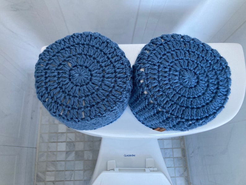 May include: Two blue crocheted toilet paper roll covers. The covers are made with a circular pattern and have a white background.