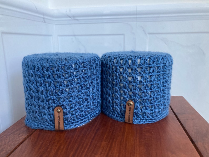 May include: Two blue crocheted toilet paper roll covers with brown leather straps and brass buckles.