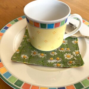 May include: A yellow and blue patterned ceramic coffee cup with a handle sits on a green floral coaster on a white plate with a colorful border.