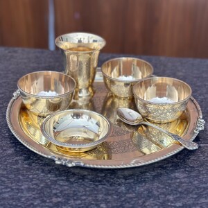 May include: A polished gold-tone metal serving set consisting of a round tray with a beaded edge, one tall tumbler, four small bowls with intricate etched patterns, and a single matching spoon. The items are arranged together on the tray.