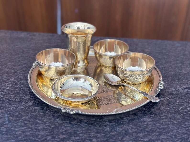 May include: A polished gold-tone metal serving set consisting of a round tray with a beaded edge, one tall tumbler, four small bowls with intricate etched patterns, and a single matching spoon. The items are arranged together on the tray.