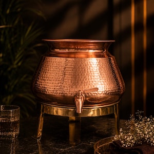 May include: A copper beverage dispenser with a hammered finish, brass tap, and stand. The dispenser has a rounded body and a flared rim. The stand has three legs. A glass of liquid and a tray with a napkin are in the background.