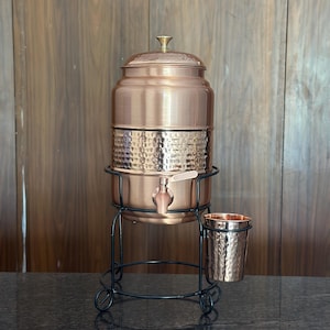 Peut inclure: Un distributeur d'eau en cuivre avec une texture martelée sur la partie inférieure, doté d'un bouton de couvercle couleur laiton et d'un robinet. Il repose sur un support en métal noir avec un porte-gobelet latéral pour une tasse assortie.