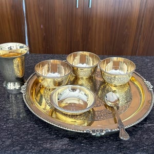 May include: A gold-toned metal serving set consisting of a round tray with an ornate border, three small bowls with textured rims, one larger bowl, a tall pedestal cup, and a single spoon. The items feature polished surfaces and intricate patterns.