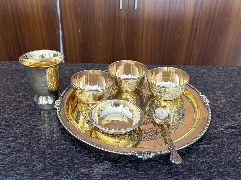 May include: A gold-toned metal serving set consisting of a round tray with an ornate border, three small bowls with textured rims, one larger bowl, a tall pedestal cup, and a single spoon. The items feature polished surfaces and intricate patterns.
