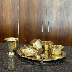 May include: A gold-colored brass dining set consisting of a round tray with a beaded edge, a tall tumbler, three small bowls of varying sizes, and a single small spoon. The items feature intricate etched floral patterns and a polished finish.