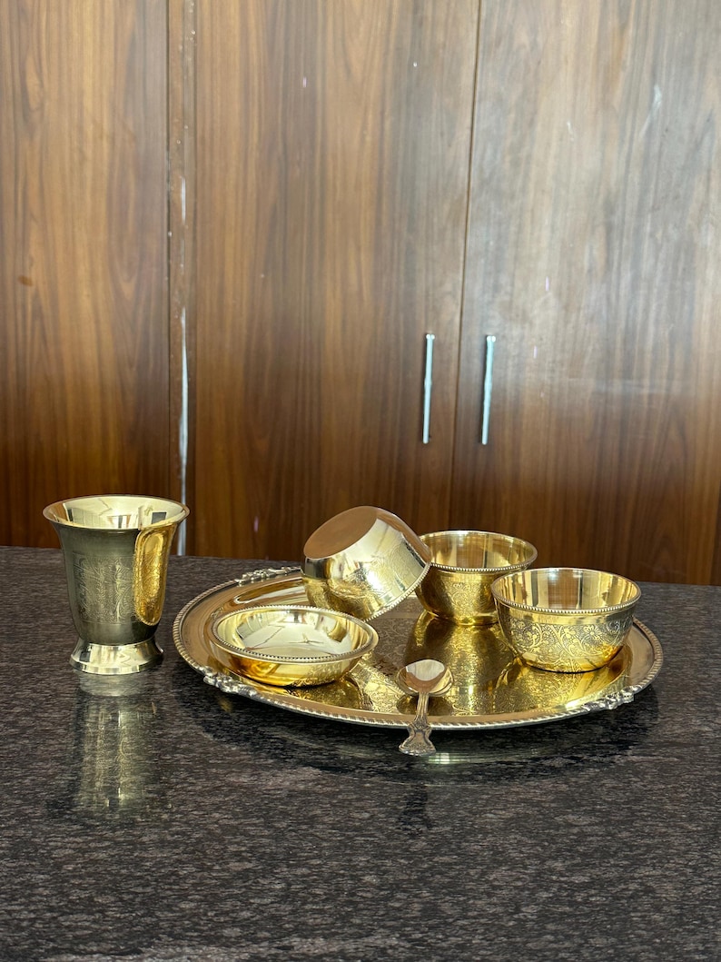 May include: A gold-colored brass dining set consisting of a round tray with a beaded edge, a tall tumbler, three small bowls of varying sizes, and a single small spoon. The items feature intricate etched floral patterns and a polished finish.