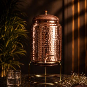 Puede incluir: Un dispensador de agua de cobre con textura martillada, un grifo y una tapa. Se encuentra sobre un soporte de metal dorado. Un vaso de agua y una pequeña bandeja con flores están en primer plano. El dispensador es decorativo.