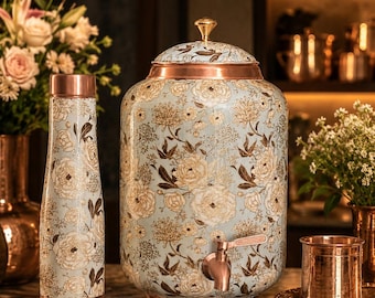 May include: A cylindrical copper water dispenser with a tap and a matching copper water bottle, both featuring a light blue background with a detailed white and gold floral pattern. The dispenser has a domed lid with a small gold knob.