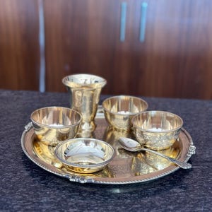 May include: A polished brass serving set consisting of a round tray with a beaded edge, a tall tumbler, three small bowls with varying textures and patterns, and a single ornate spoon. The items feature a reflective gold-toned metallic finish.