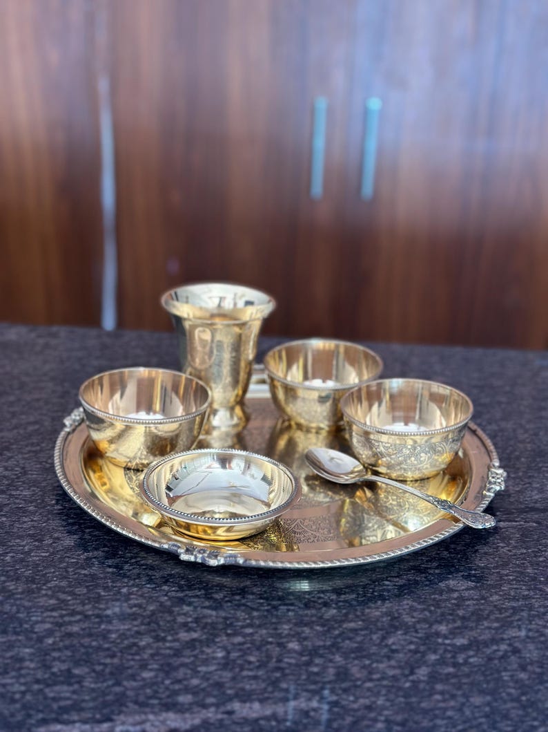 May include: A polished brass serving set consisting of a round tray with a beaded edge, a tall tumbler, three small bowls with varying textures and patterns, and a single ornate spoon. The items feature a reflective gold-toned metallic finish.