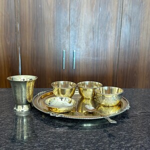 May include: A gold-toned metal set consisting of a tall tumbler, three small bowls, a shallow dish, and a spoon arranged on a round serving tray. Each piece features intricate engraved patterns and a polished finish with a beaded rim detail.