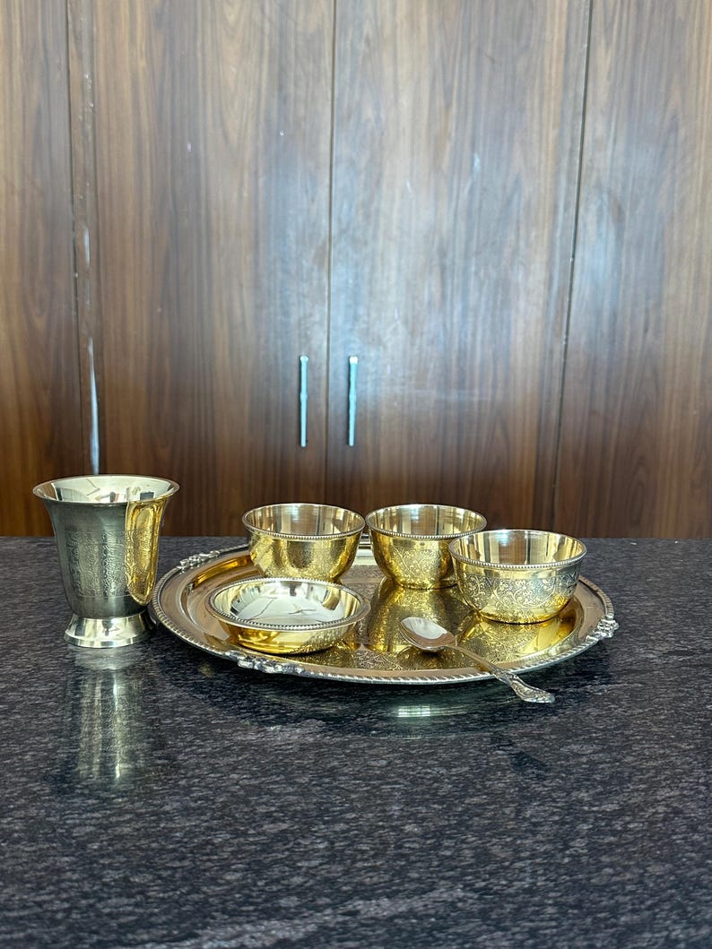May include: A gold-toned metal set consisting of a tall tumbler, three small bowls, a shallow dish, and a spoon arranged on a round serving tray. Each piece features intricate engraved patterns and a polished finish with a beaded rim detail.