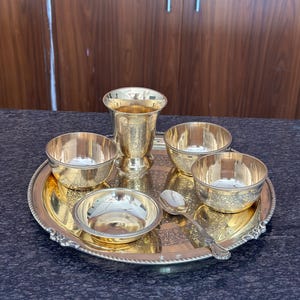 May include: A gold-colored metal serving set consisting of a round tray with an ornate border, a tall tumbler, three small bowls, and a single spoon. Each piece features intricate floral engravings and a polished finish, arranged together as a set.