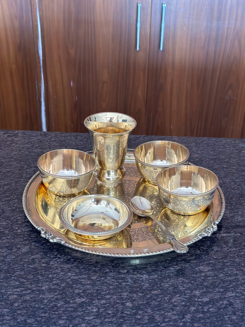 May include: A gold-colored metal serving set consisting of a round tray with an ornate border, a tall tumbler, three small bowls, and a single spoon. Each piece features intricate floral engravings and a polished finish, arranged together as a set.