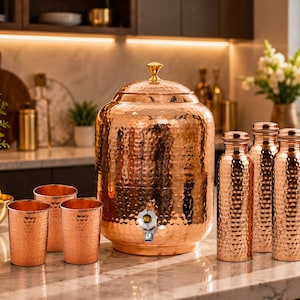 May include: A large hammered copper water dispenser with a gold-toned lid knob and a silver-toned spigot, accompanied by three matching hammered copper tumblers and three matching hammered copper water bottles with silver-toned caps.