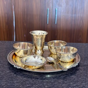 May include: A polished brass dining set consisting of a large round serving tray with a beaded edge, a tall tumbler, three small bowls with intricate floral engravings, and a single matching spoon, all featuring a reflective metallic finish.