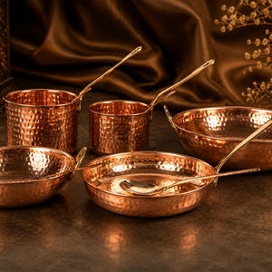 May include: A selection of copper cookware with a hammered finish. The set includes bowls, pots, and serving spoons. The copper pieces have a warm, reddish-brown colour and are arranged on a dark surface.
